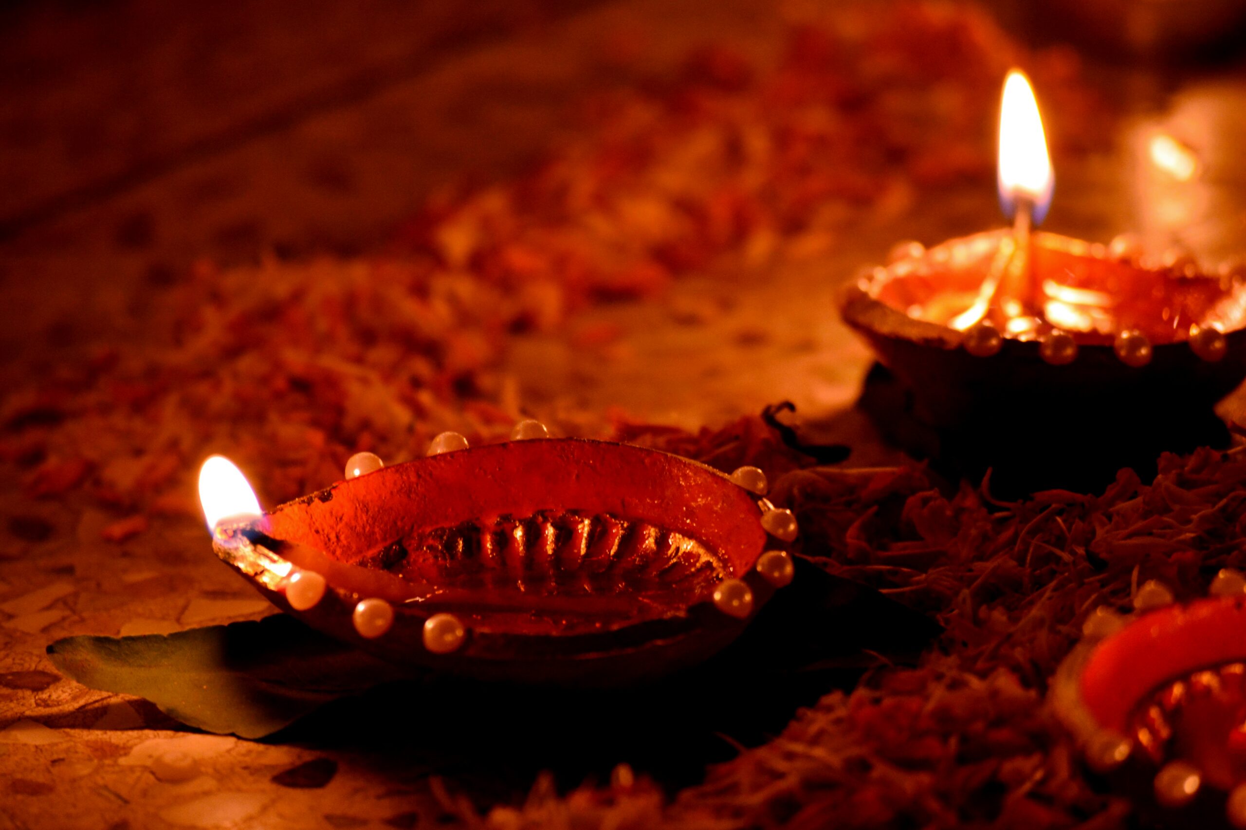 Glowing oil lamps (diyas) with bright flames sit on a textured surface, casting a warm orange light and creating a festive, peaceful mood.